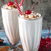 Two tall glasses of vanilla milkshake topped with whipped cream, chocolate shavings, and a cherry, each with a red-and-white striped straw. A bowl of cherries is visible in the background.