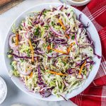 A bowl of coleslaw with shredded green and purple cabbage, carrots, and sliced red onion, seasoned with black pepper, sits on a white surface next to a red-and-white checkered napkin—the perfect side for sautéed mushrooms for steak.