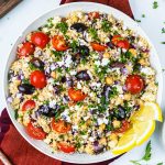 A bowl of Mediterranean quinoa salad with cherry tomatoes, kalamata olives, chickpeas, feta cheese, chopped parsley, red onion, and lemon wedges on the side.