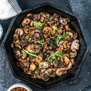A black skillet filled with Sautéed Mushrooms for Steak, garnished with fresh rosemary and chopped herbs, sits on a dark surface alongside a napkin and small bowls.