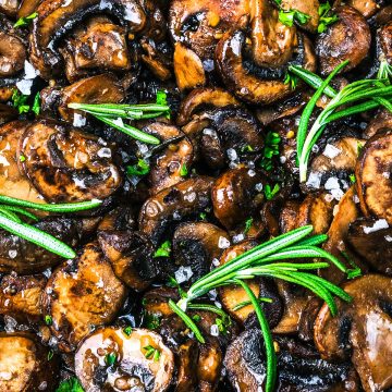 Sliced sautéed mushrooms for steak, garnished with fresh rosemary and sprinkled with coarse sea salt. The mushrooms are browned and glistening, giving a savory and appetizing appearance.