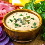 A bowl of creamy sauce topped with chopped pickles and herbs, surrounded by sliced tomatoes, leaf lettuce, sliced red onions, and cheese on a wooden surface.