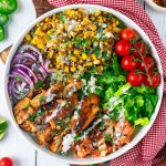 A bowl filled with grilled chicken, charred corn, sliced red onions, cherry tomatoes on the vine, chopped lettuce, salsa, cilantro, and drizzled white sauce, arranged in colorful sections on a white table.