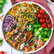 A bowl filled with grilled chicken, charred corn, sliced red onions, cherry tomatoes on the vine, chopped lettuce, salsa, cilantro, and drizzled white sauce, arranged in colorful sections on a white table.