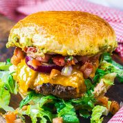 A close-up of a juicy cheeseburger with melted cheddar, lettuce, red onion, tomato salsa, and a golden toasted bun, sitting on a wooden surface with a red checkered cloth in the background.