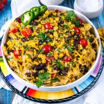A bowl of taco pasta salad with farfalle, ground beef, black beans, cherry tomatoes, shredded cheese, jalapeño slices, cilantro, and crushed tortilla chips, served on a colorful plate.