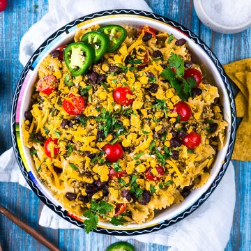 A colorful bowl of pasta salad with cherry tomatoes, black beans, shredded cheese, jalapeño slices, cilantro, and crushed corn chips on top, set on a blue wooden table with a white napkin and tortilla chips nearby.