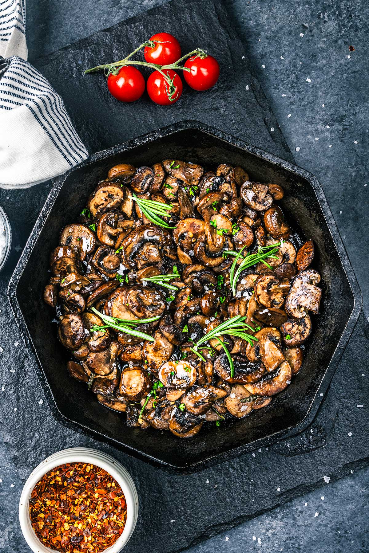 A black skillet filled with Sautéed Mushrooms for Steak, garnished with fresh herbs, sits on a slate surface. Nearby are vine tomatoes, a small bowl of chili flakes, and a striped cloth napkin.