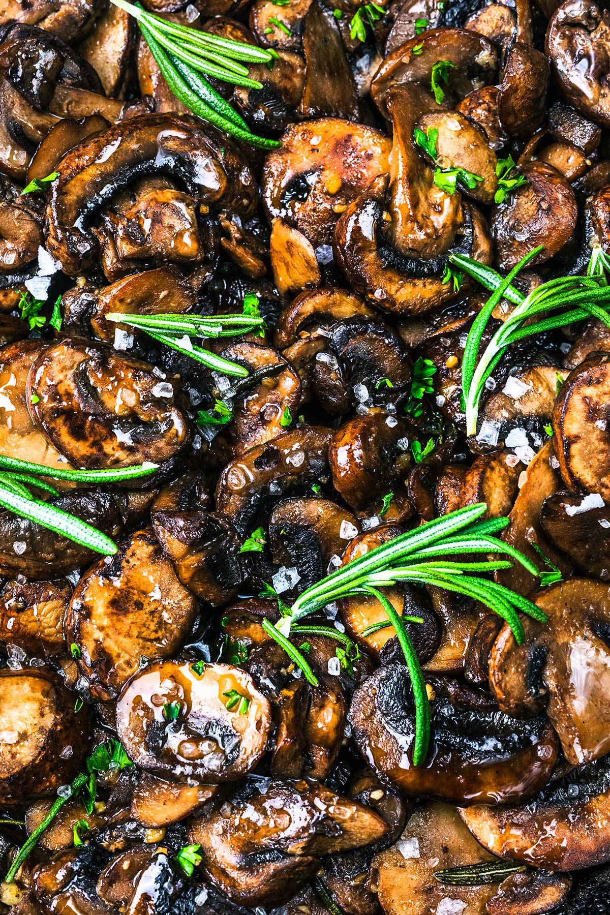 Close-up of Sautéed Mushrooms for Steak, garnished with fresh rosemary sprigs, chopped chives, and coarse sea salt, glistening with oil and seasonings.