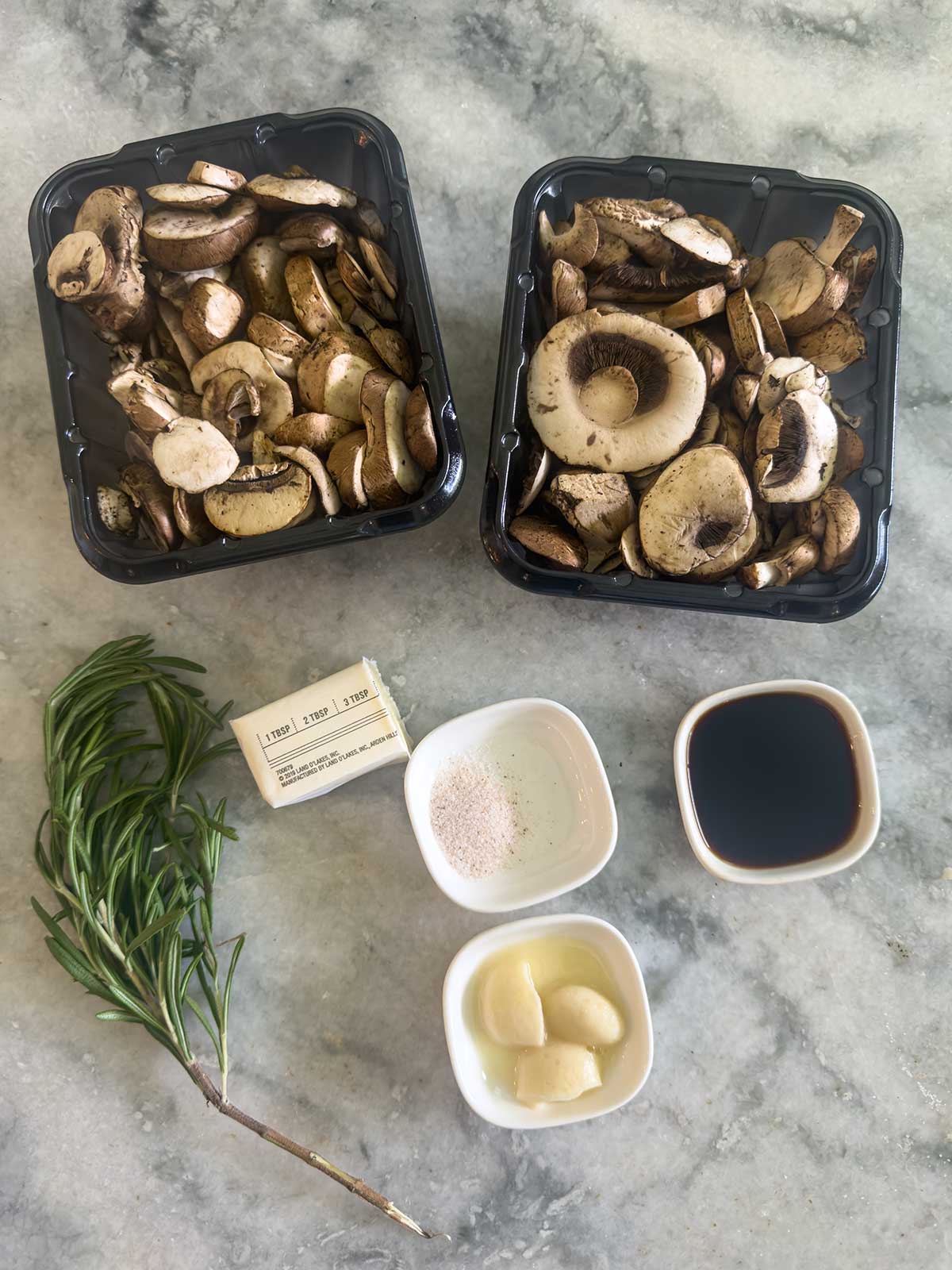 Two black containers of sliced mushrooms, a sprig of rosemary, two sticks of butter, three garlic cloves, and bowls of salt, balsamic vinegar, and soy sauce arranged on marble—perfect ingredients for flavorful Sautéed Mushrooms for Steak.