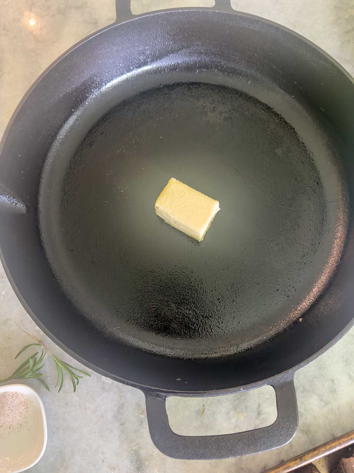 A chunk of butter sits in the center of a black cast iron skillet on a light-colored countertop, ready to melt for sautéed mushrooms for steak, with a small bowl of salt and a sprig of rosemary nearby.