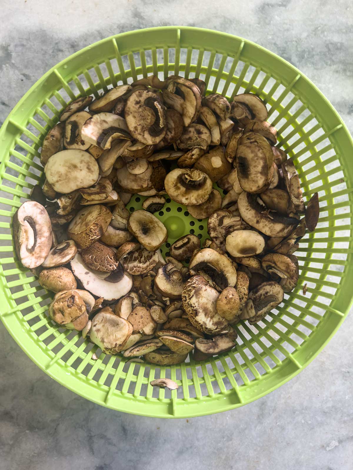 A green plastic colander filled with sliced mushrooms, perfect for sautéed mushrooms for steak, sits on a gray marble countertop.