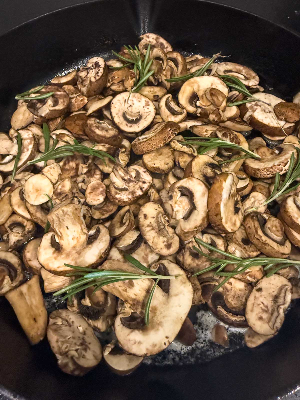 Sautéed mushrooms for steak and sprigs of fresh rosemary cook in a black skillet, with visible melted butter or oil at the bottom.
