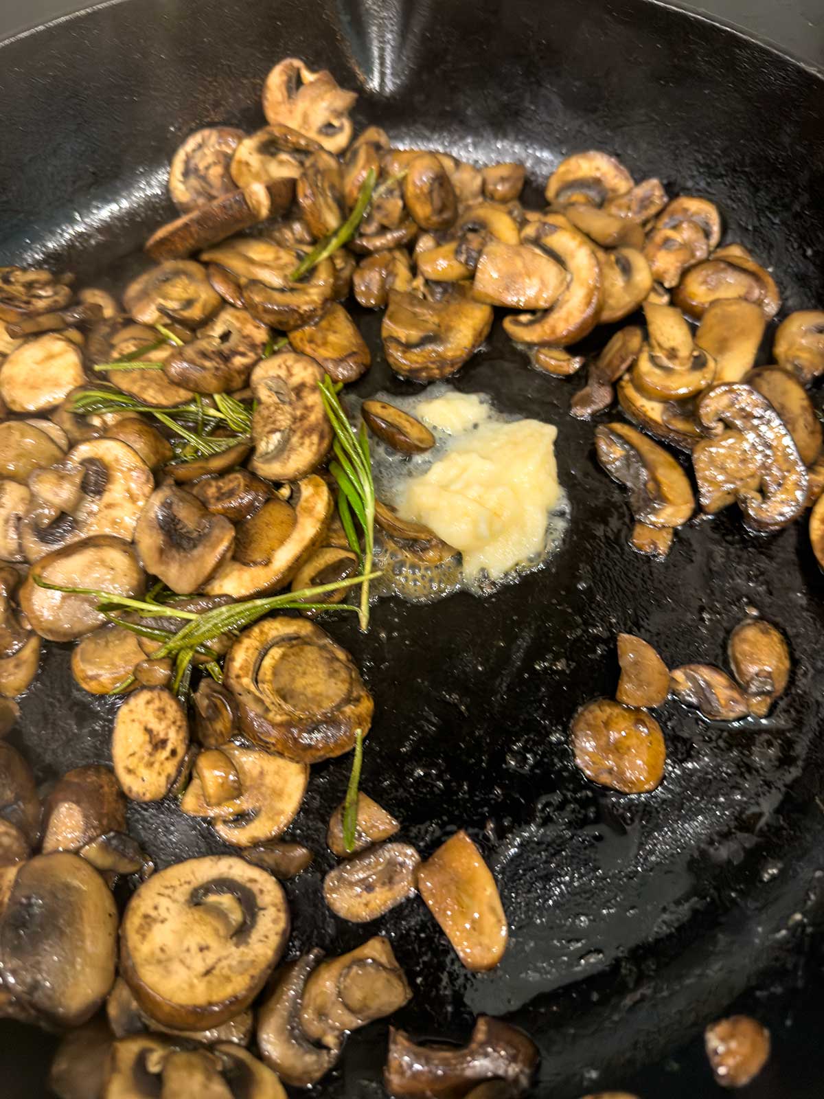 Sautéed Mushrooms for Steak: Sliced mushrooms and sprigs of rosemary are sizzling in a cast iron skillet, with a dollop of minced garlic melting in the center—perfect as a savory topping for your favorite steak.