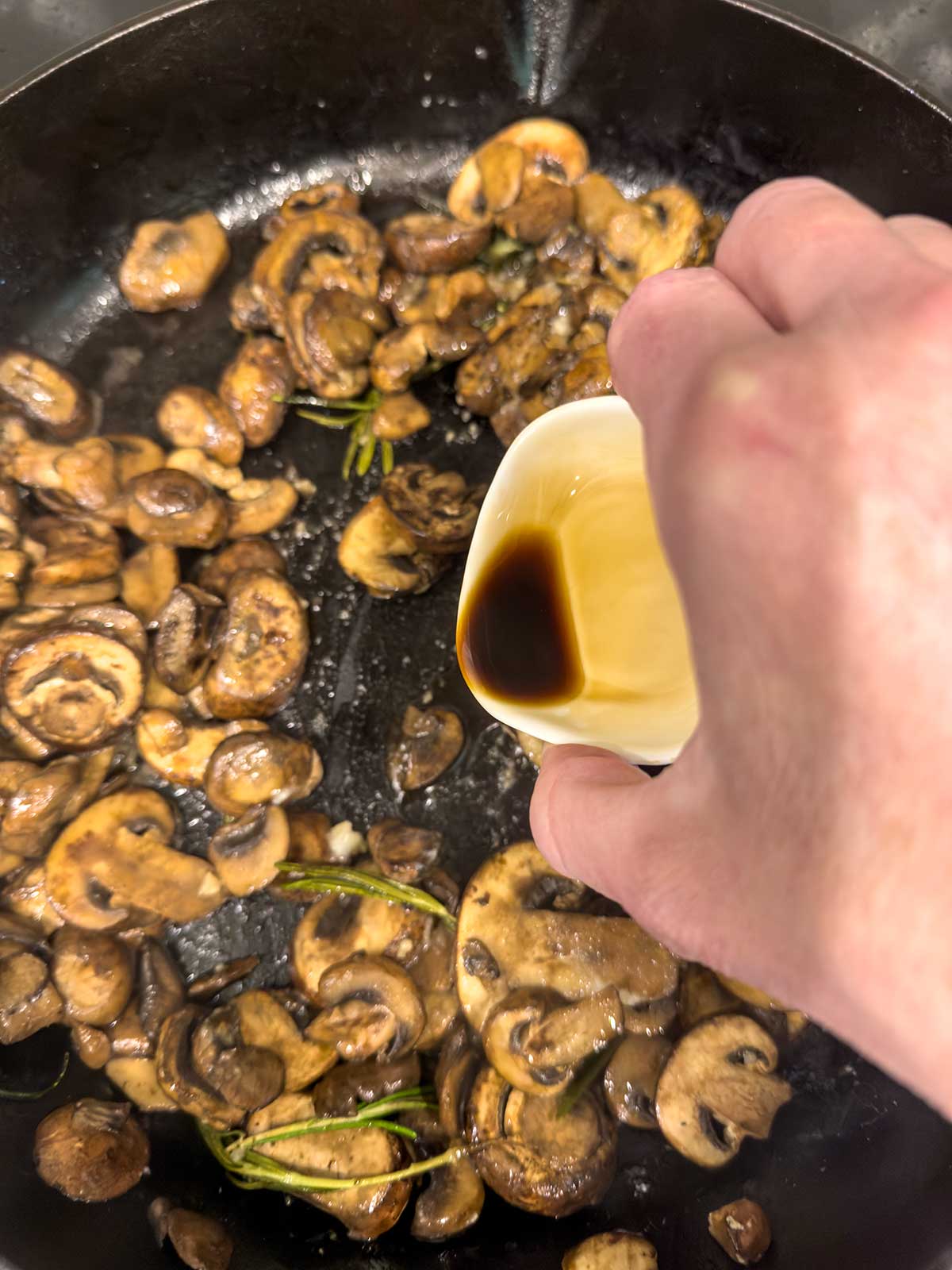 A hand pours soy sauce from a small white dish onto sautéed mushrooms for steak cooking in a black skillet, with some rosemary sprigs nestled among the mushrooms.
