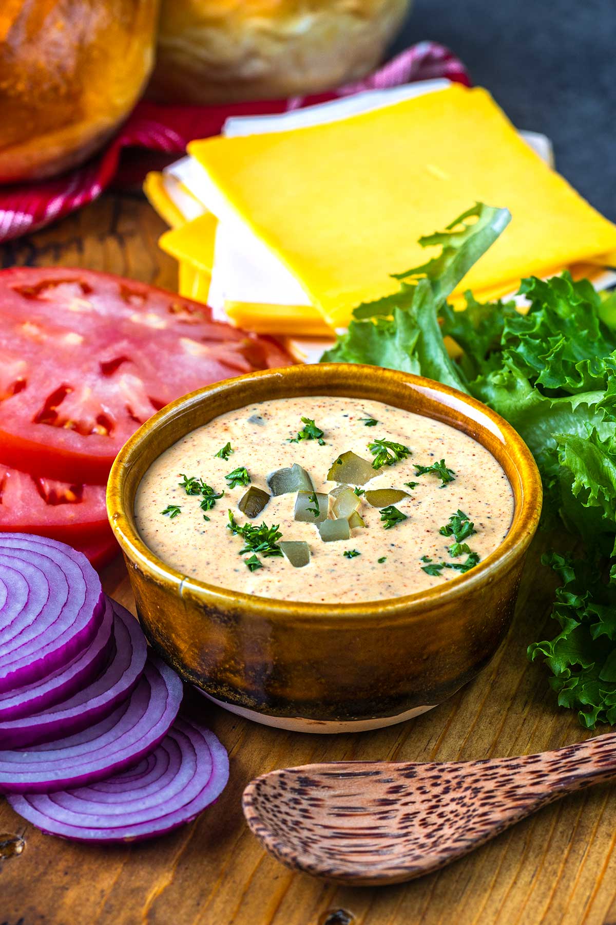 A small bowl of creamy dip topped with chopped pickles and herbs is surrounded by sliced tomatoes, red onions, lettuce, and stacked cheese slices on a wooden table with a wooden spoon nearby.