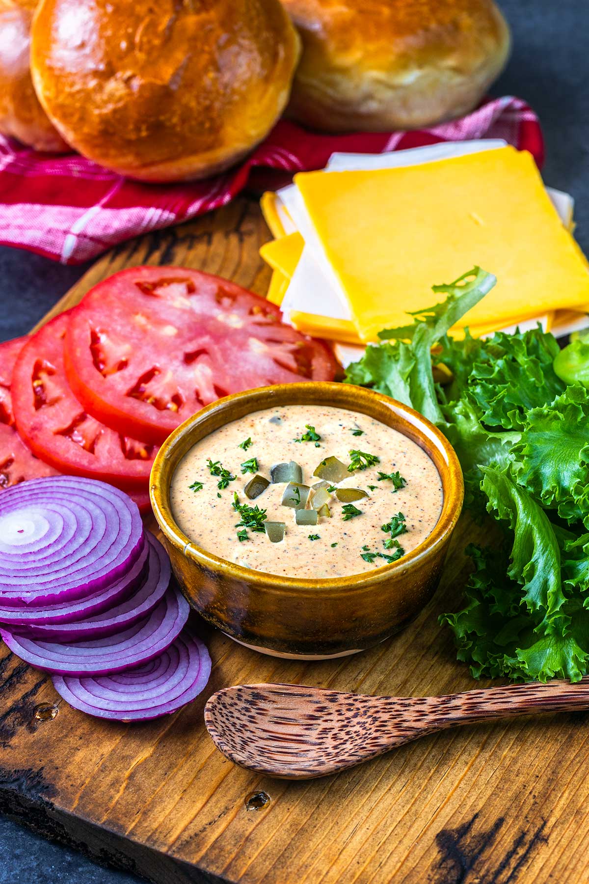 A wooden board with sliced red onion, tomato, leafy lettuce, cheddar cheese slices, a bowl of creamy dip topped with herbs and pickles, a wooden spoon, and round bread rolls in the background.
