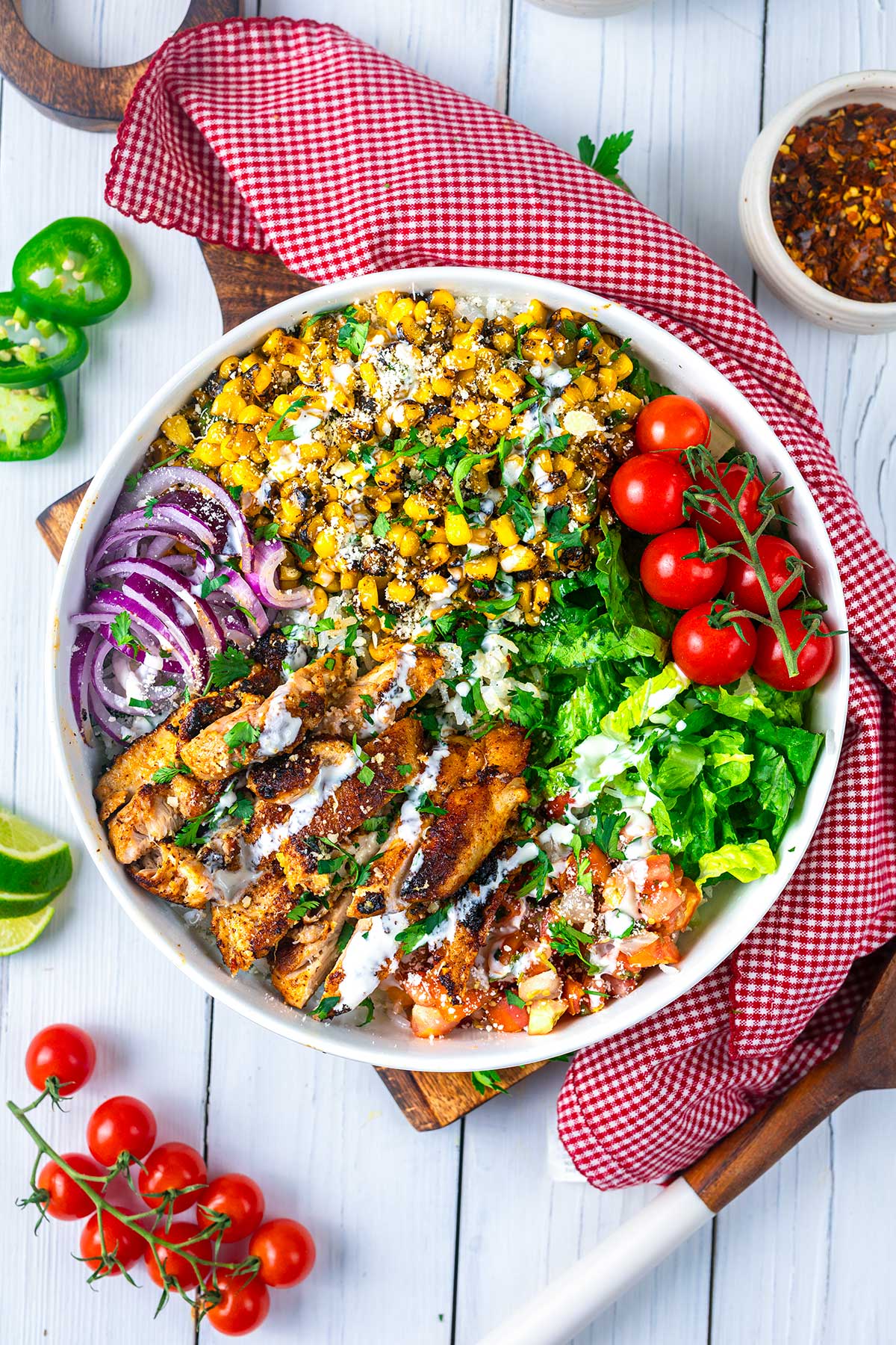 A colorful salad bowl with grilled chicken slices, grilled corn, cherry tomatoes, red onion, lettuce, and pico de gallo, drizzled with white sauce. The bowl is on a red checkered napkin, with fresh ingredients around it.
