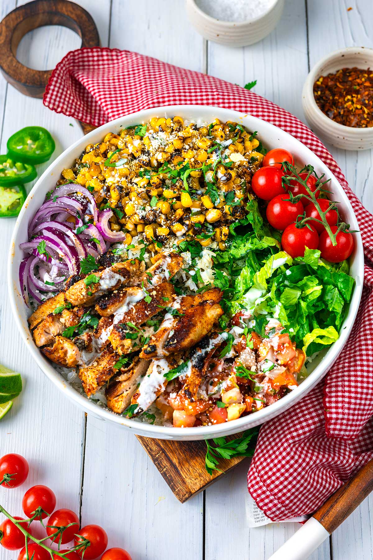 A bowl filled with grilled chicken, corn, cherry tomatoes, red onion, romaine lettuce, salsa, and crumbled cheese, garnished with herbs and dressing. The bowl sits on a white wooden table with a red checkered napkin.