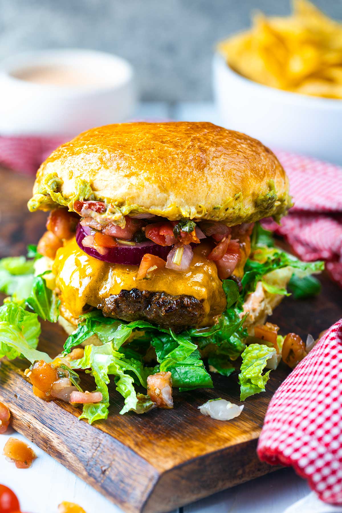 A juicy cheeseburger with lettuce, cheddar cheese, red onion, and salsa on a wooden board, with tortilla chips and a dipping sauce in the background.