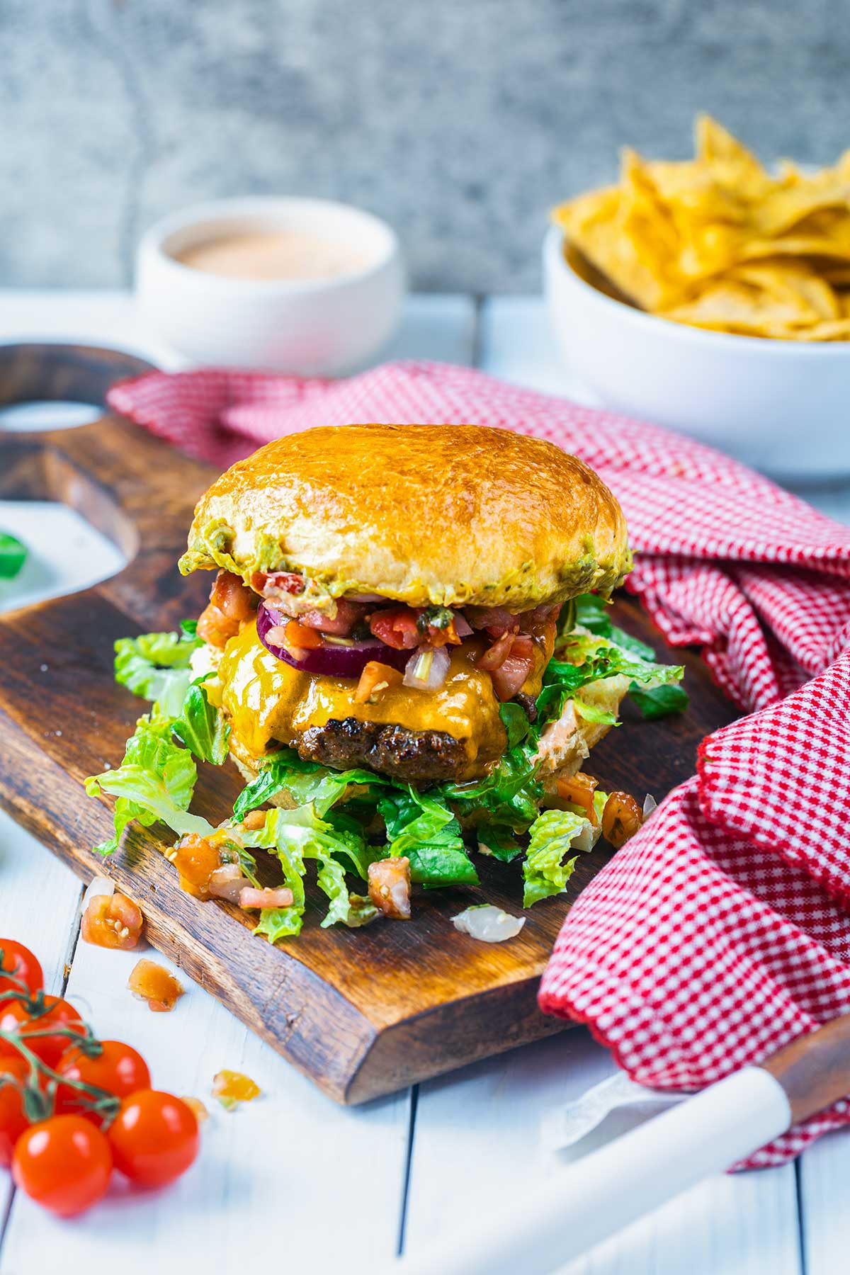 A cheeseburger topped with lettuce, diced tomatoes, sauce, and a brioche bun sits on a wooden board. Nearby are cherry tomatoes, a bowl of tortilla chips, and a red checkered cloth.
