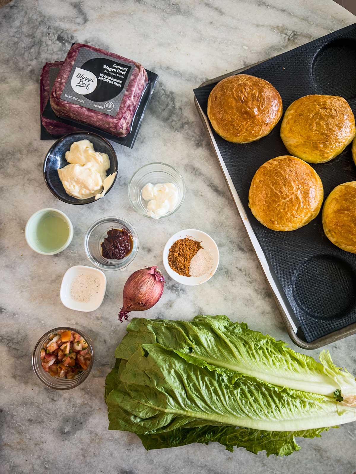 Overhead view of burger ingredients on a marble surface: ground beef, four brioche buns, romaine lettuce, sliced shallot, aioli, mayonnaise, spices, sauce, pickled vegetables—perfect alongside a fresh taco pasta salad and a small bowl of juice or vinegar.