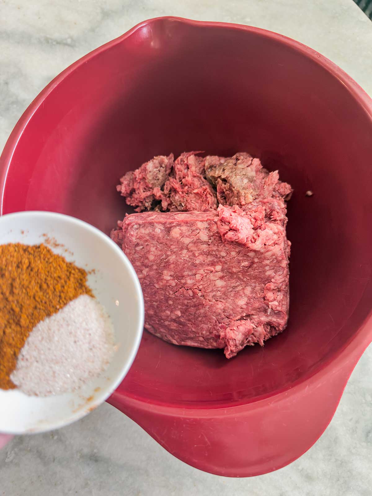 A red mixing bowl with raw ground beef sits on a countertop. In the foreground, a hand holds a small white bowl of spices and salt, ready to be added—perfect for making flavorful taco pasta salad.