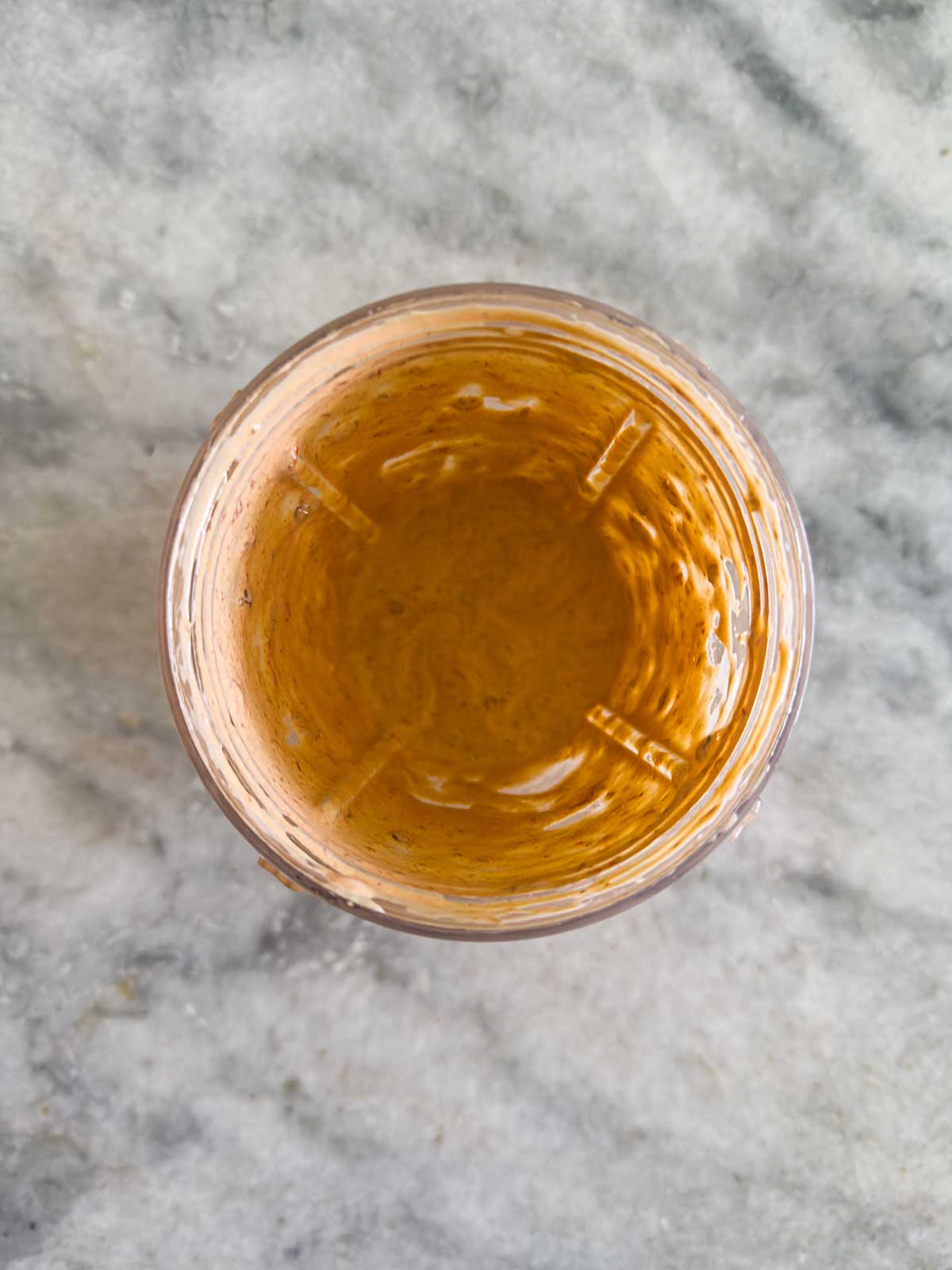 A top-down view of a blender cup containing a small amount of smooth, light brown sauce or dressing for taco pasta salad, set on a light gray marble surface.