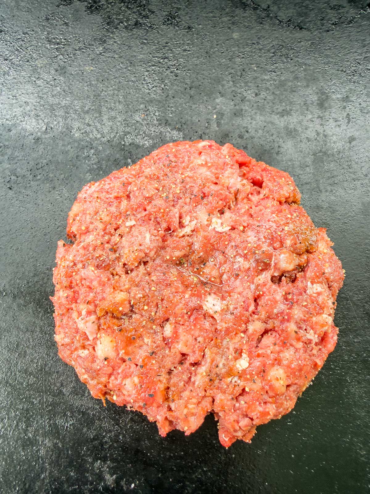 A raw ground beef patty, seasoned and shaped into a round form, sits on a dark, flat surface ready to be cooked—perfect for pairing with a zesty taco pasta salad.