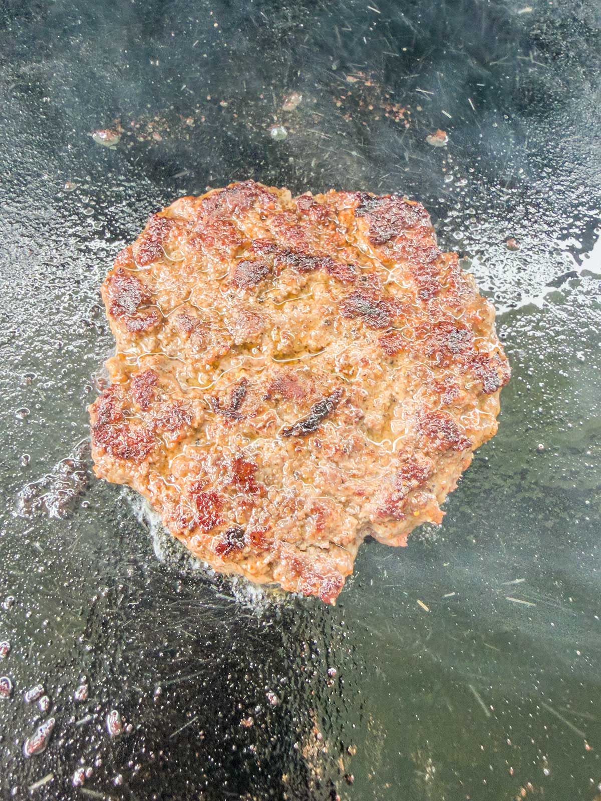 A single beef burger patty with a crispy, browned exterior cooks on a flat, oily griddle surface, surrounded by sizzling grease—right next to a bowl of vibrant taco pasta salad.