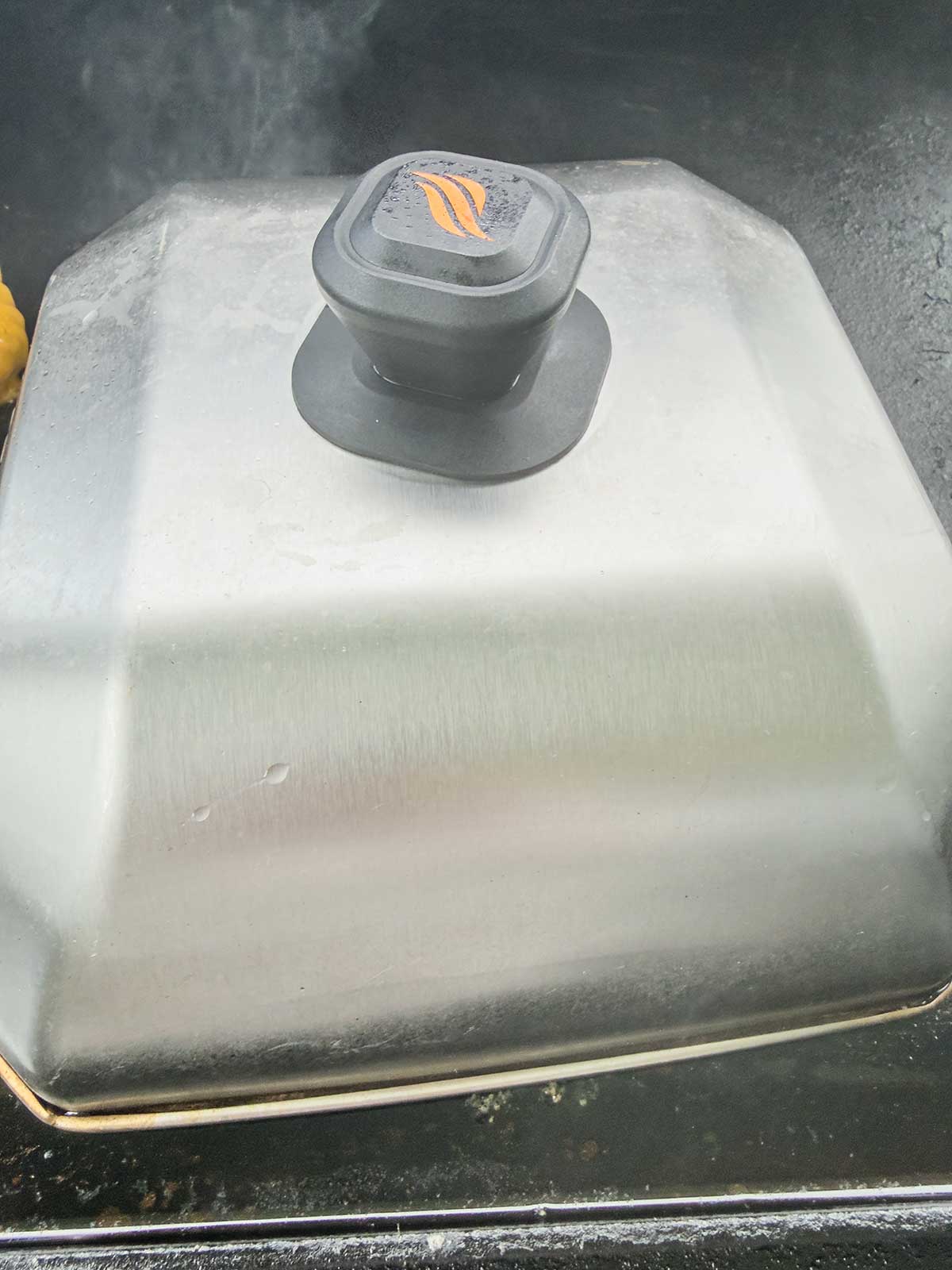 A close-up of a metal grilling dome cover with a black handle, sitting on a grill surface with steam visible around the edges—perfect for sealing in flavor before serving dishes like taco pasta salad.