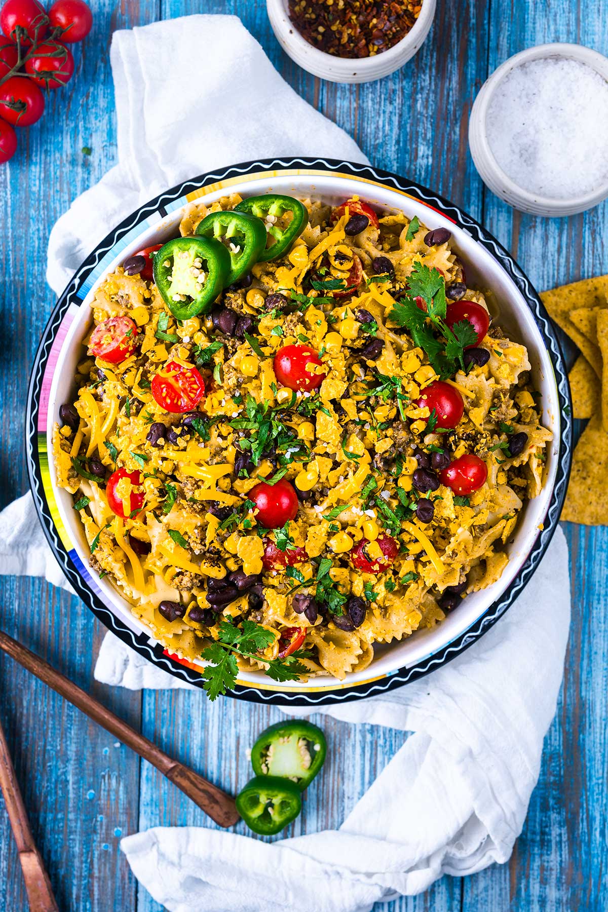 A bowl of nachos topped with melted cheese, black beans, cherry tomatoes, sliced jalapeños, fresh cilantro, and crumbled tortilla chips, set on a blue wooden table with a white napkin and garnishes nearby.