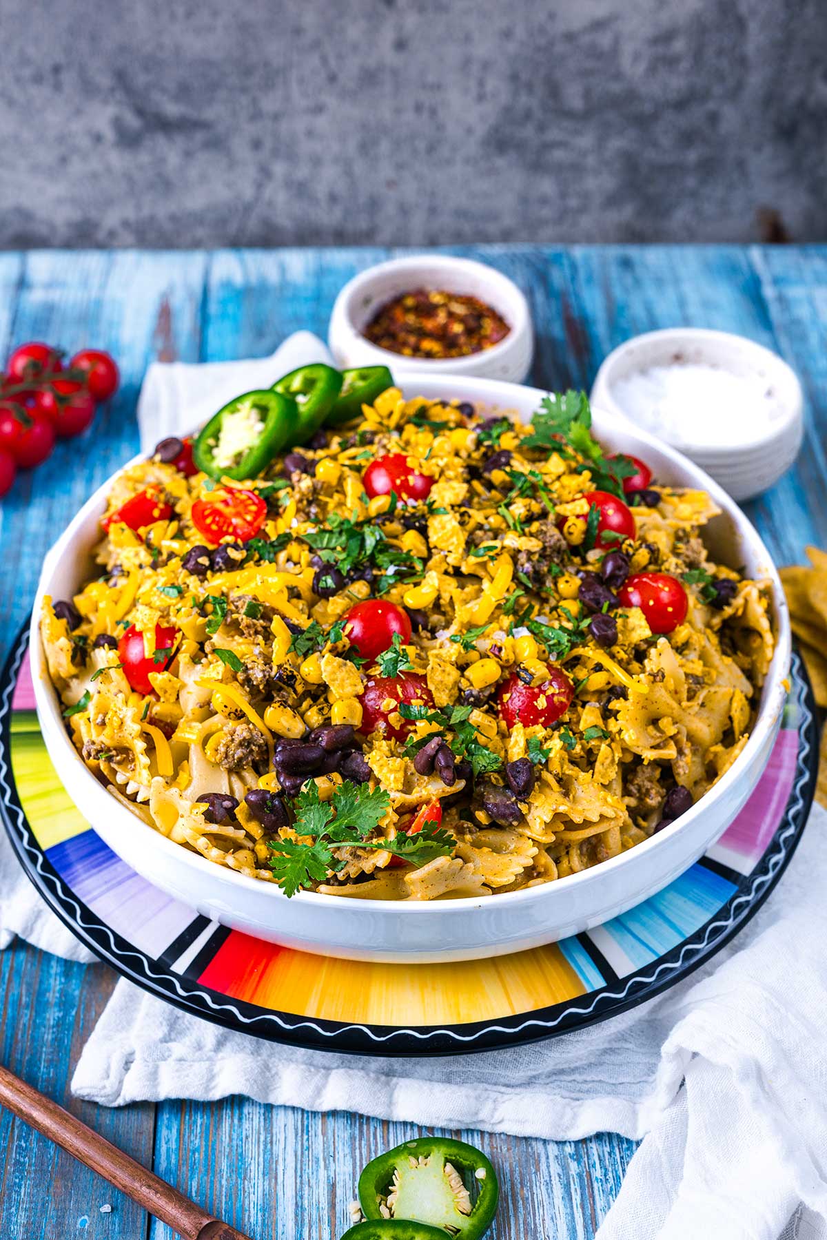 A colorful bowl of taco pasta salad with bowtie pasta, black beans, cherry tomatoes, crushed tortilla chips, cheese, jalapeños, and cilantro, placed on a vibrant plate with bowls of seasoning and salt in the background.
