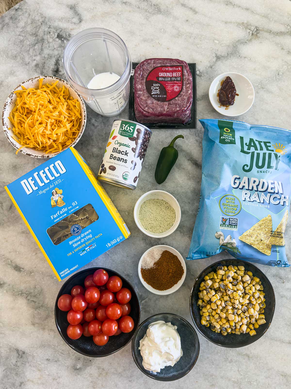 Ingredients for taco pasta salad displayed on a marble surface, including shredded cheddar, farfalle pasta, black beans, cherry tomatoes, sour cream, ground beef, corn, jalapeño, spices, ranch chips, chipotle pepper, and milk in a blender cup.