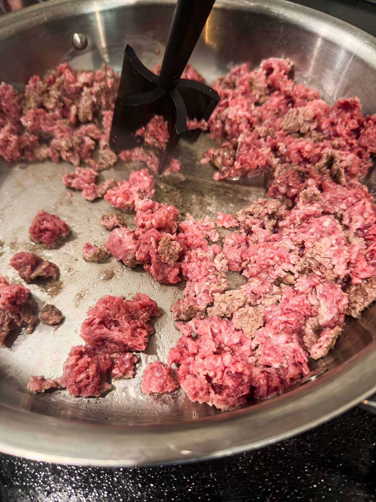 Ground beef is being browned in a stainless steel skillet for a taco pasta salad, with a black meat chopper breaking up the meat as it cooks. Some beef is still pink while other pieces are turning brown.
