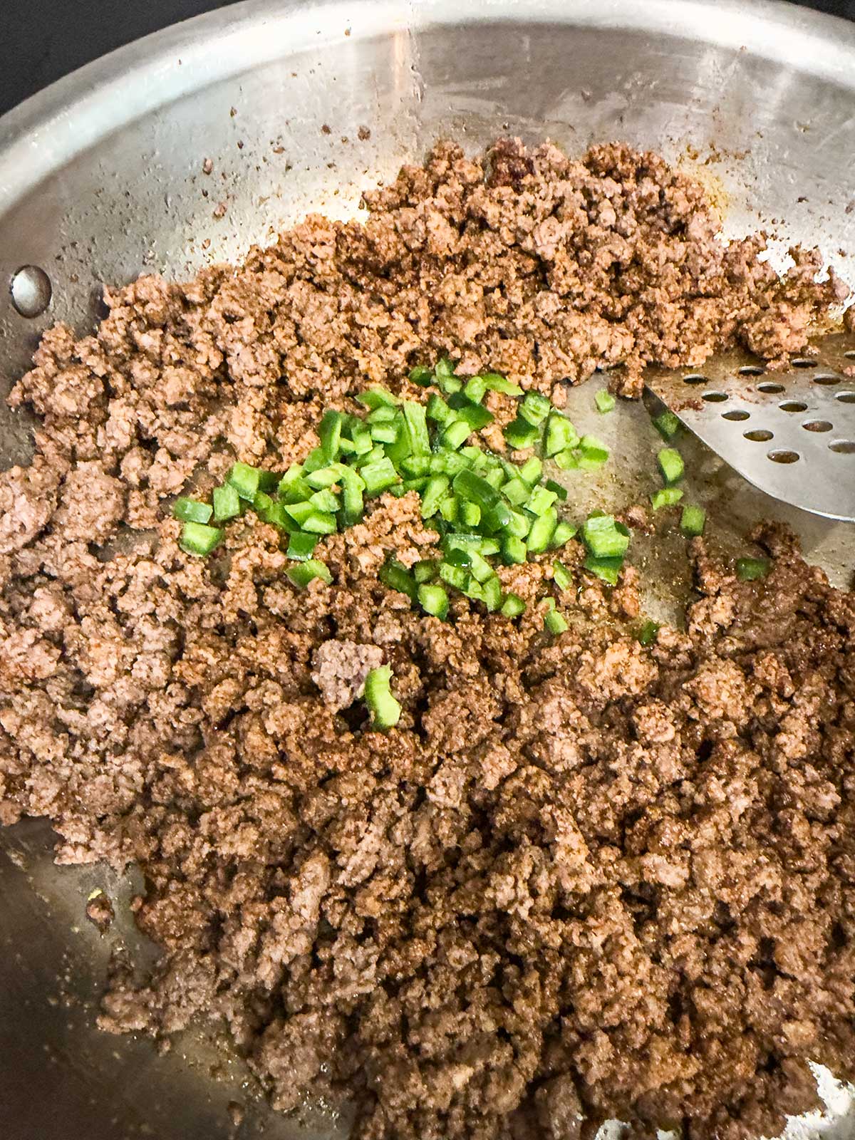 Ground beef cooking in a stainless steel skillet, with chopped green peppers added in the center for a flavorful taco pasta salad, and a slotted metal spatula resting on the side of the pan.