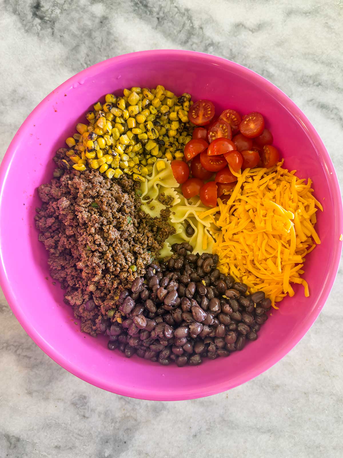 A pink bowl on a marble surface holds a vibrant taco pasta salad, with ground beef, black beans, shredded cheddar, halved cherry tomatoes, corn, and cooked noodles arranged separately around the bowl.