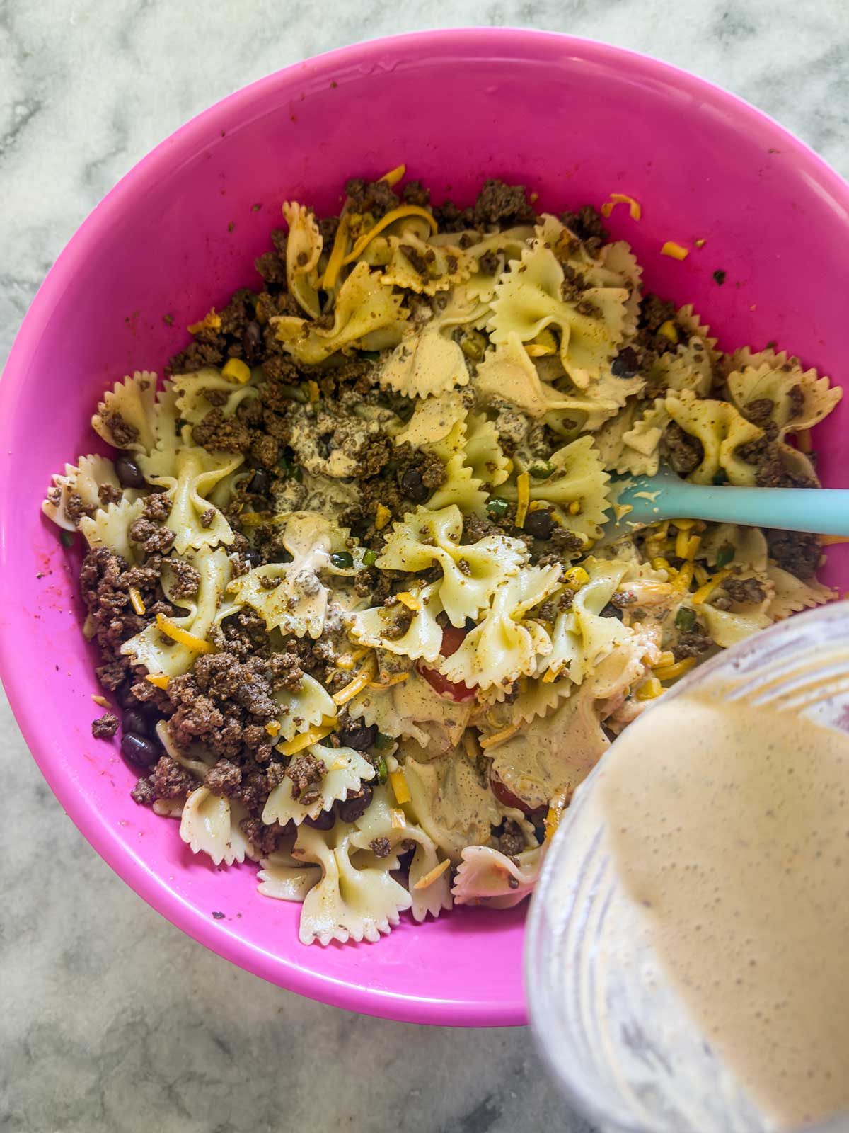 A pink bowl filled with taco pasta salad—bowtie pasta, ground beef, black beans, shredded cheese, and spices—while a creamy dressing is poured from a glass container into the mixture. A blue spoon rests in the bowl.