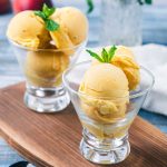 Two glass cups filled with scoops of peach sorbet each garnished with a sprig of mint, placed on a wooden board with a blurred background.