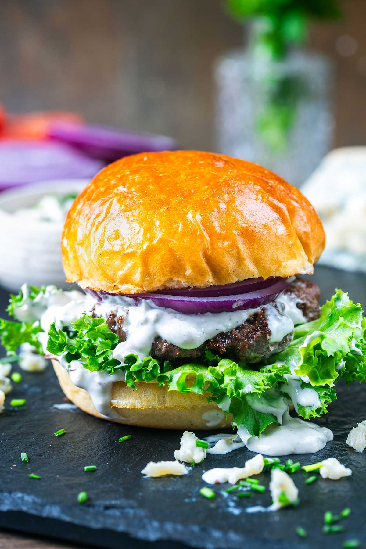 A juicy blue cheese burger topped with leafy lettuce, red onion slices, and creamy white sauce on a shiny brioche bun, served on a black slate with chopped chives and crumbled cheese scattered around.