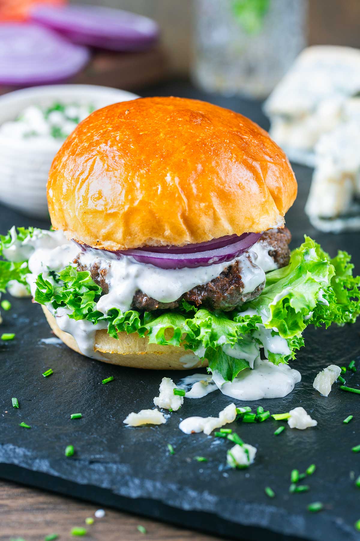 A blue cheese burger with a shiny brioche bun, leafy lettuce, beef patty, creamy white sauce, and red onion slices sits on a dark slate, garnished with chives and crumbled cheese.