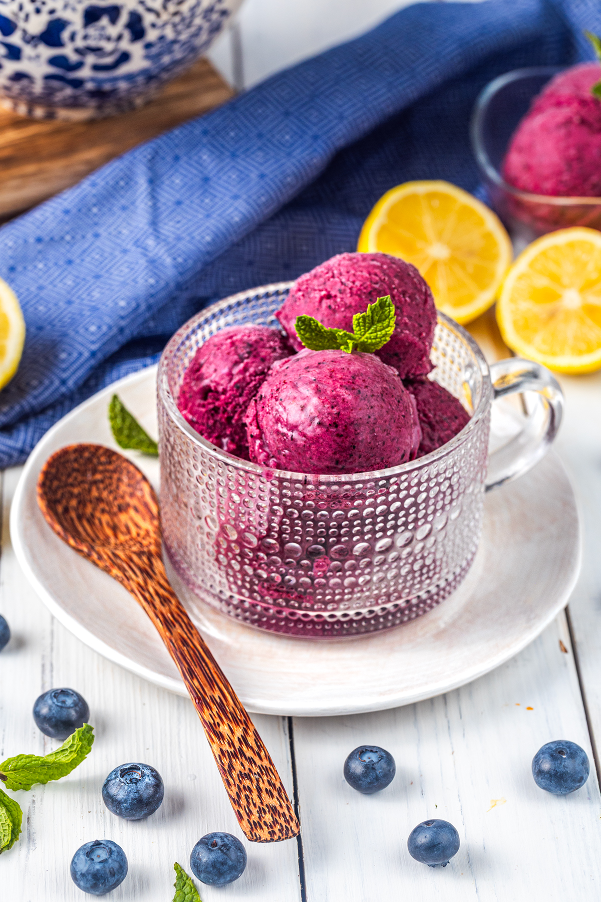 A glass cup filled with three scoops of purple berry sorbet, garnished with mint, sits on a white plate with a wooden spoon. Blueberries, lemon halves, and a blue cloth are in the background.