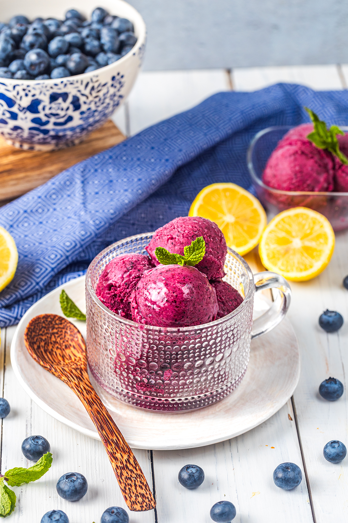 A glass cup with scoops of blueberry sorbet, garnished with mint, sits on a white plate with a wooden spoon. Blueberries and lemon halves are scattered nearby, with a bowl of blueberries and blue cloth in the background.