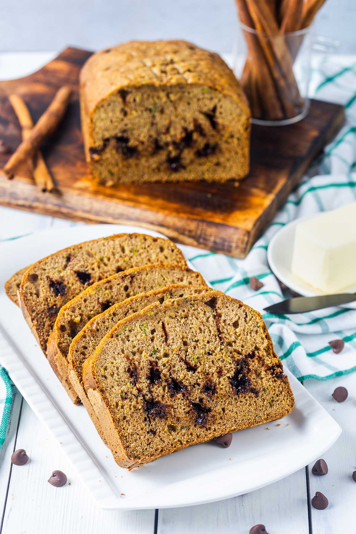 Sliced zucchini bread with chocolate chips on a white plate, with a loaf and cinnamon sticks on a wooden board in the background, and a dish of butter nearby.