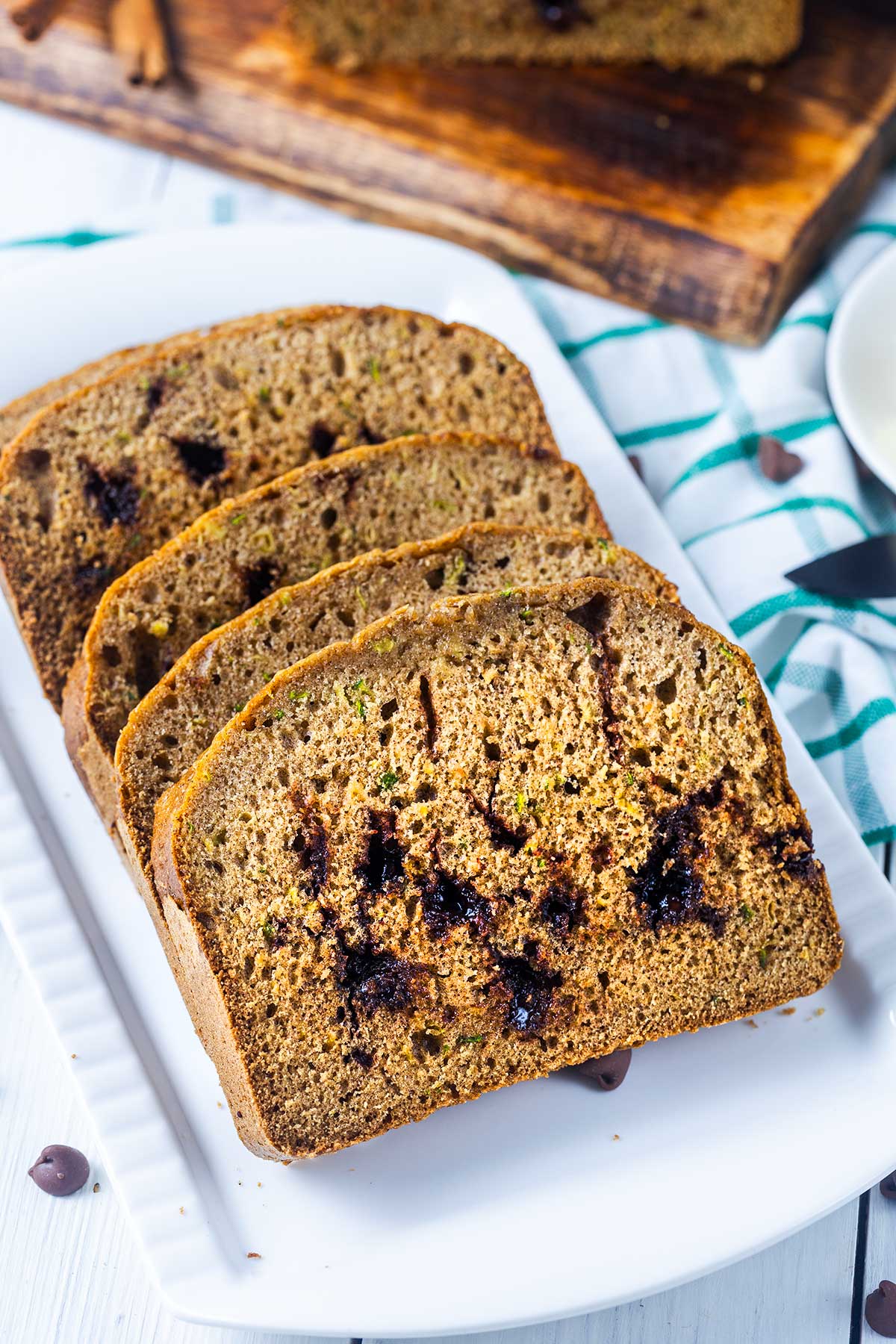 Four slices of chocolate chip zucchini bread are arranged in a row on a white plate, with a green-striped towel and a wooden cutting board in the background.