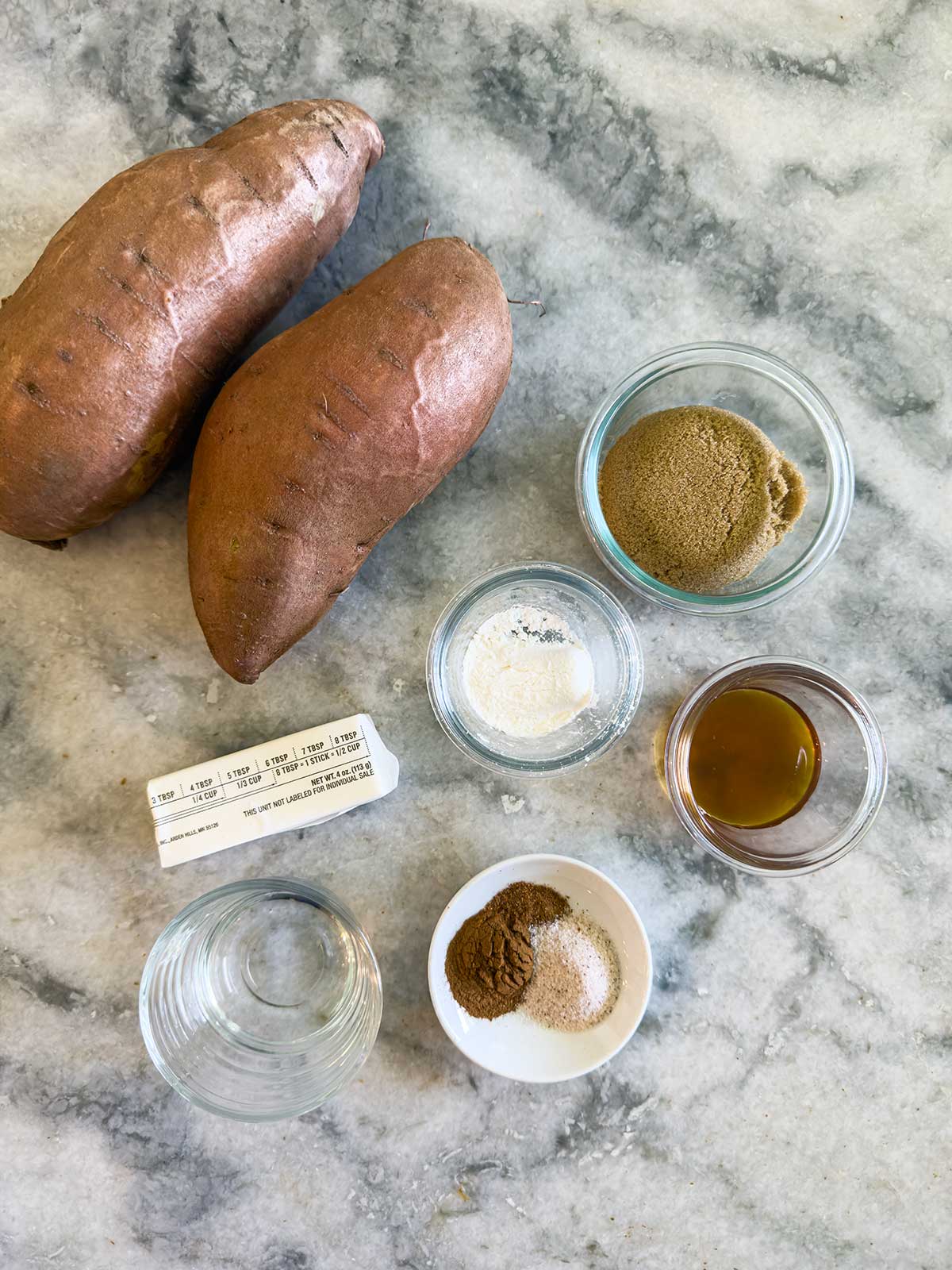 Two whole sweet potatoes sit next to small bowls of brown sugar, melted butter, vanilla extract, water, and a dish with cinnamon and nutmeg on a marble countertop.