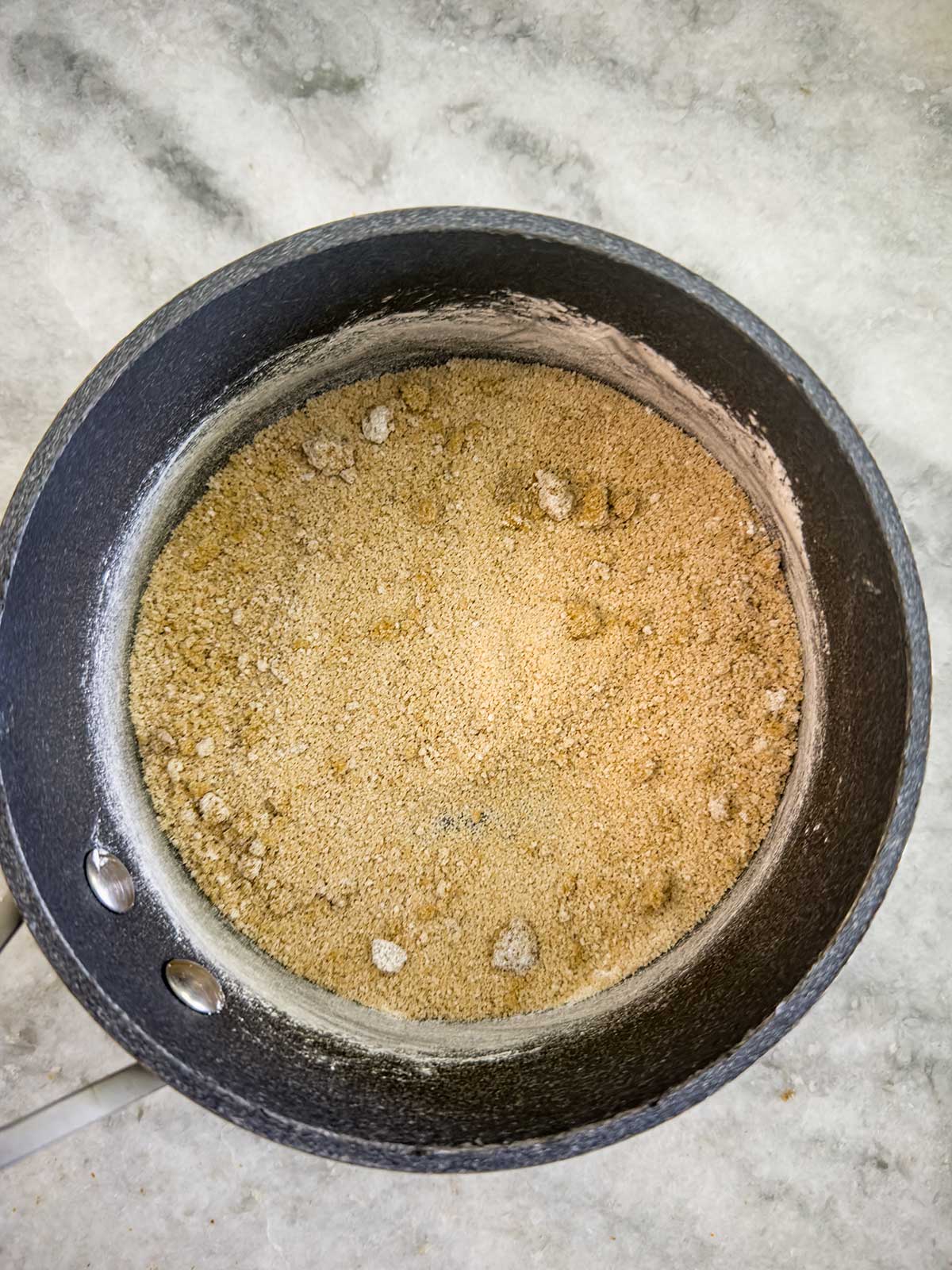 A black saucepan filled with dry, light brown crumbs, likely brown sugar and possibly other dry ingredients, sits on a light-colored marble countertop.