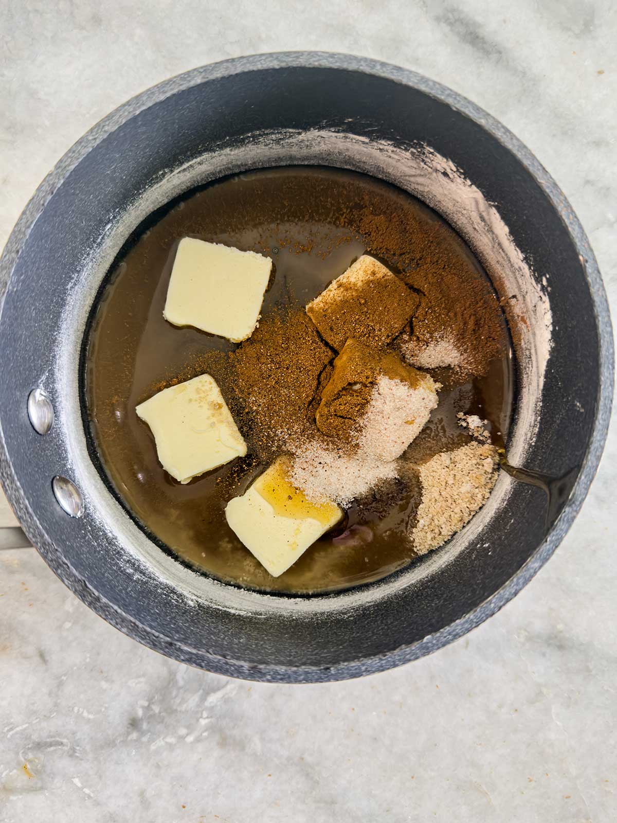 A saucepan with butter slices, brown sugar, white sugar, spices, and flour on a marble surface, ready to be mixed together.