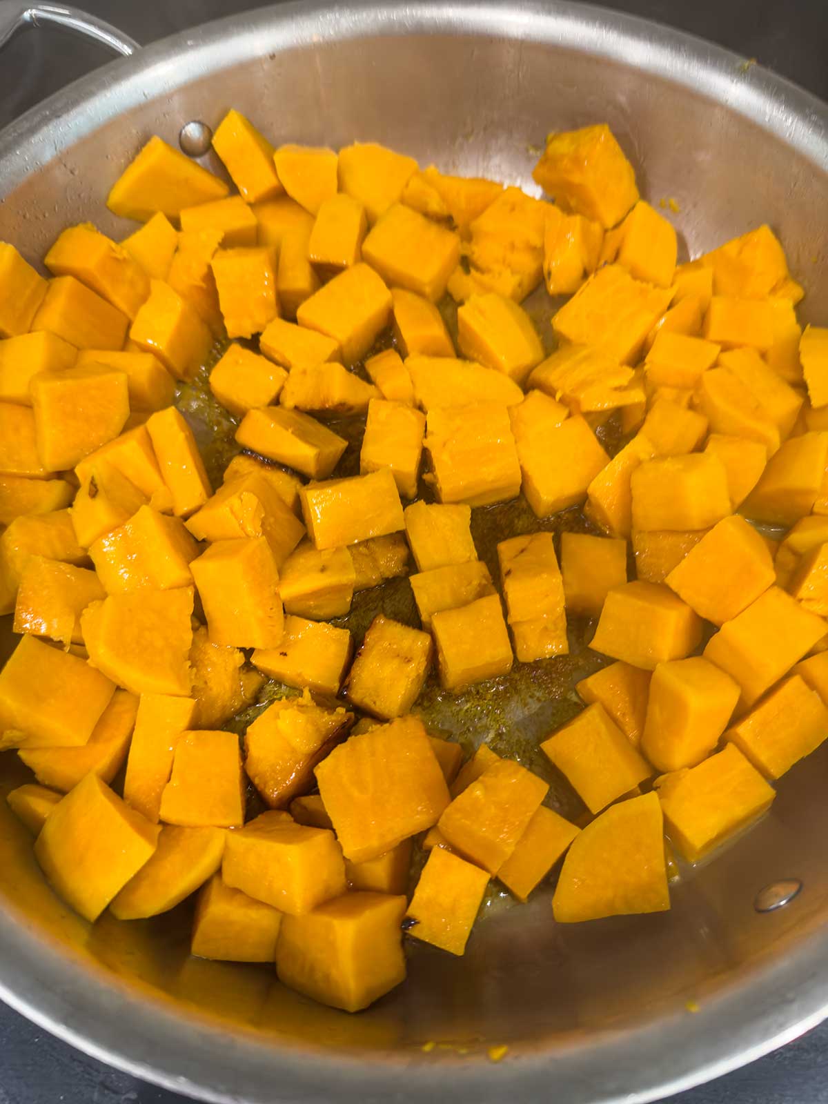 A close-up of cubed orange squash or sweet potato pieces cooking in a large stainless steel pan with some oil.