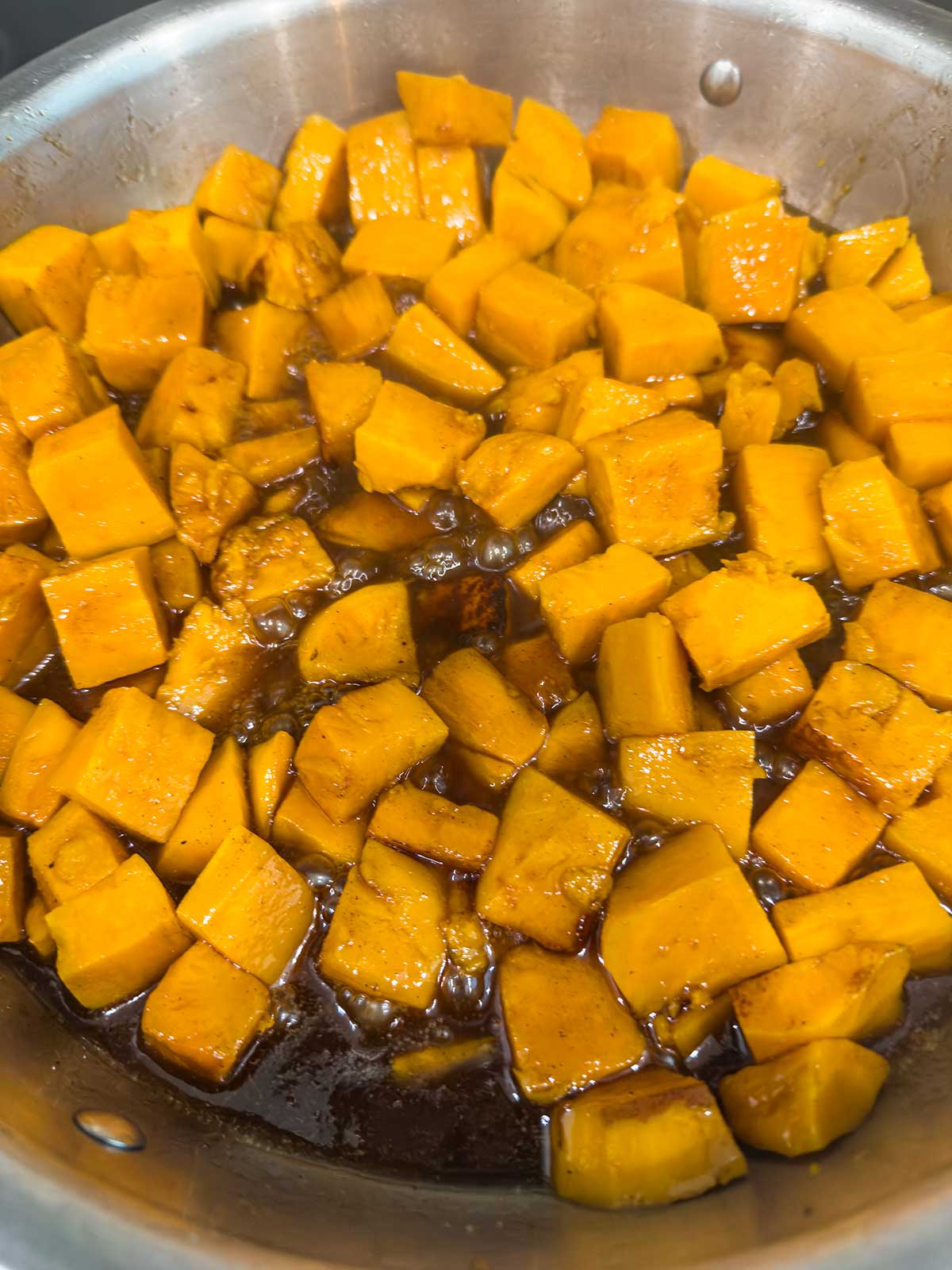 Cubed pieces of orange sweet potatoes simmering in a pot with a brown liquid, likely syrup or sauce, partially covering the pieces.
